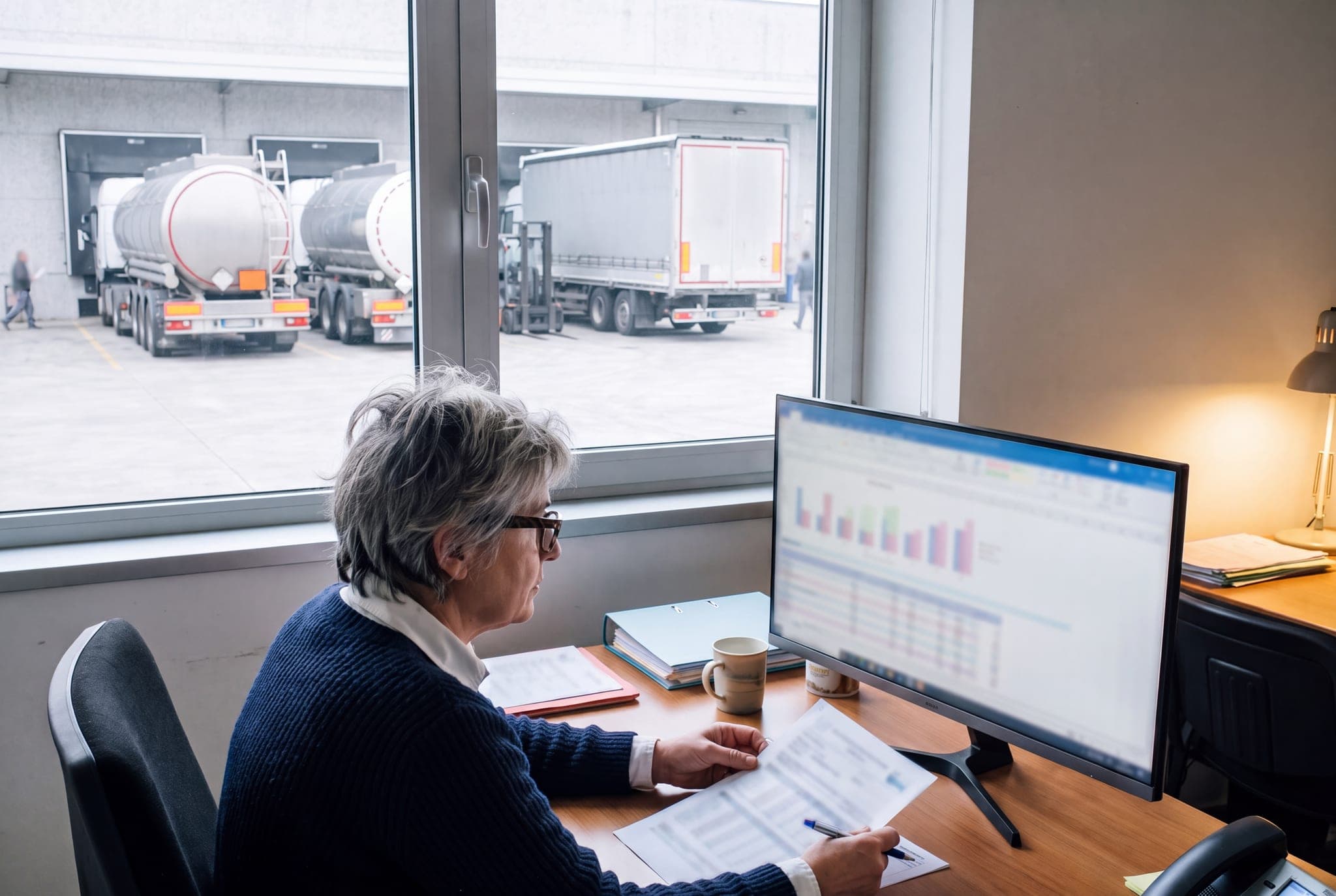 A logistics manager compares a printed transport invoice with the information on her screen. Through the window, two tankers and a curtain-sided lorry from different carriers can be seen in a loading yard.